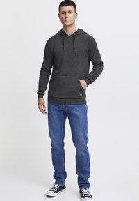 Gray hooded sweatshirt with front pocket, ribbed cuffs, and hem. Paired with blue denim jeans and black sneakers, standing against a plain background.