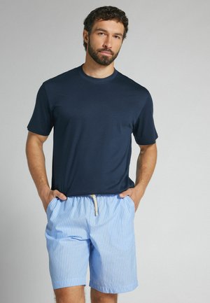 Mann mit Bart, der ein marineblaues T-Shirt und hellblaue gestreifte Shorts trägt, mit Händen in den Taschen vor einem schlichten Hintergrund.