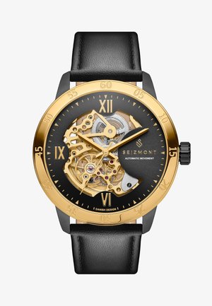 Luurankoautomaatikello kultainen ja musta värimaailma. Varustettu mustalla nahkarannekkeella, kultaisilla tuntimerkeillä ja monimutkaisella mekanismilla.