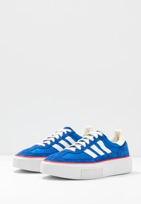 Blue suede sneakers with white stripes, a thick white platform sole, flat laces, and red accent around the sole. Textured upper and smooth lining.