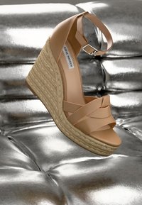 Sandale compensée nude avec des sangles entrecroisées, une plateforme en jute tissé et une bride de cheville. Matériau lisse sur une surface métallique réfléchissante.