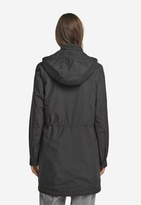 Svart lång parka jacka med huva, åtdragen midja och knappar vid ärmsluten. Slät tyg med matt yta, inga synliga mönster eller logotyper.