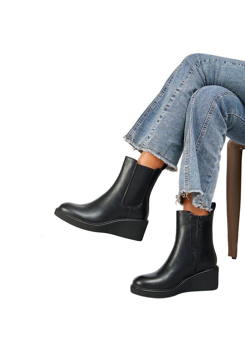 Bottes basses en cuir noir avec semelles compensées et panneaux latéraux élastiques, portées avec un jean à ourlet effiloché, assise sur une chaise.