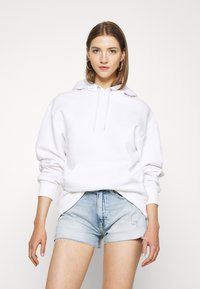 Felpa bianca in cotone con tasca a marsupio, cappuccio con cordino e polsini a coste, abbinata a pantaloni shorts in denim chiaro strappati.