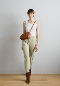 Bolso cruzado de cuero marrón, camiseta sin mangas blanca, pantalones verdes claros y botas de ante marrón hasta el tobillo. Modelo caminando frente a un fondo neutro.