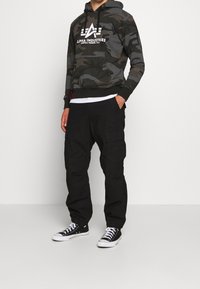 Kamouflage hoodie med vit logga, svarta cargobyxor och svarta Converse-skor. Mjuk tyg, avslappnad passform och fickdetaljer finns.