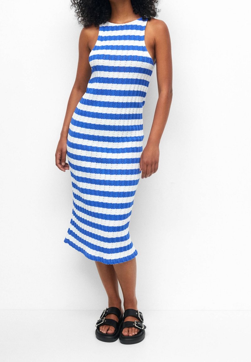 Femme portant une robe midi sans manches à rayures horizontales bleues et blanches avec des sandales noires à boucle, debout sur fond blanc.