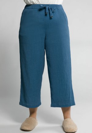 Pantalon large texturé bleu, court, avec taille à cordon, porté avec des chaussons beiges pelucheux et un haut blanc.