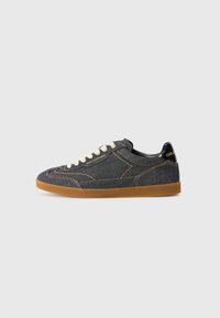 Sapatilhas de denim com um cabedal azul escuro, costuras laranja em contraste e uma sola de borracha crepe. Apresentam atacadores brancos e um detalhe em preto no calcanhar.