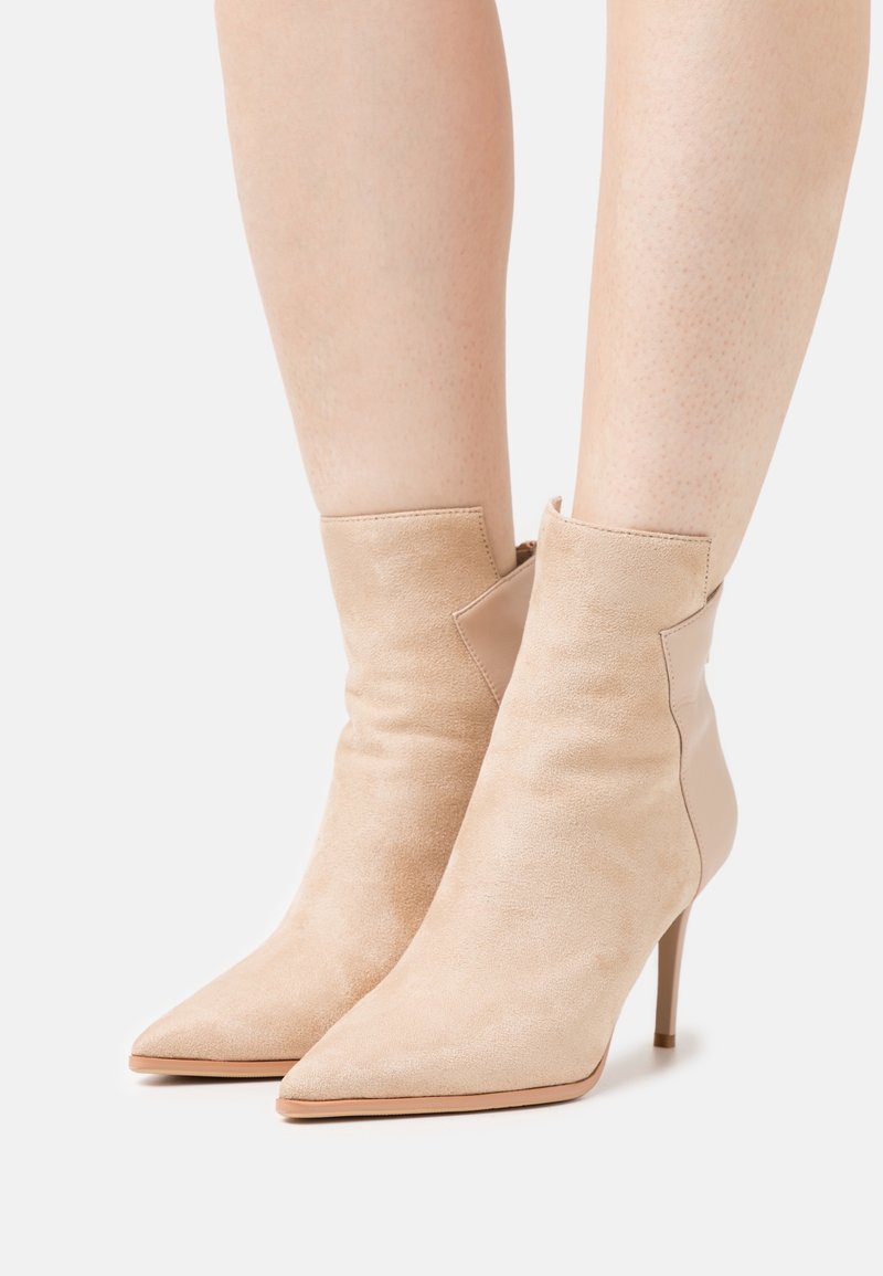 Bottes chevilles en daim beige avec bouts pointus et talons hauts fins, portées sur des jambes nues, sur un fond uni.