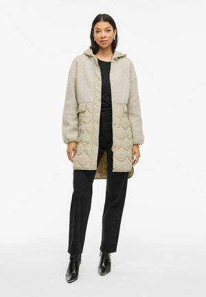 Femme portant un manteau beige matelassé en polaire avec capuche, un haut noir, un pantalon noir et des bottines noires, debout devant un fond blanc.