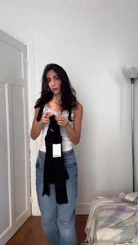 Schwarzes Oberteil, gehalten von einer Person, die ein weißes Tanktop und ausgewaschene blaue Jeans trägt. Weißes Etikett am Kleidungsstück sichtbar. Im Hintergrund weiche Polstermöbel.