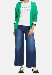 Žena v zeleném kardiganu přes bílém tričku s nápisem "SUMMER SEA YOU IN", širokých modrých džínech a bílých sand álech, stojící na bílém pozadí.