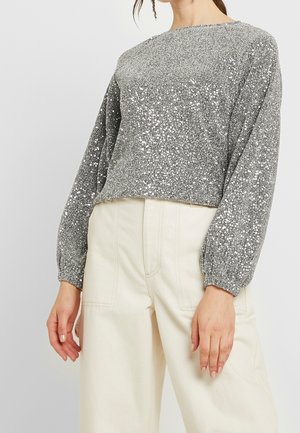 Vrouw draagt een zilverkleurige top met lange mouwen en pofmouwen, versierd met pailletten, en roomkleurige, wijde broek met hoge taille.