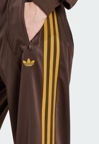 Pantaloni sportivi maronii cu trei dungi galbene pe latură. Au un buzunar cu fermoar și un logo galben brodat aproape de talie.