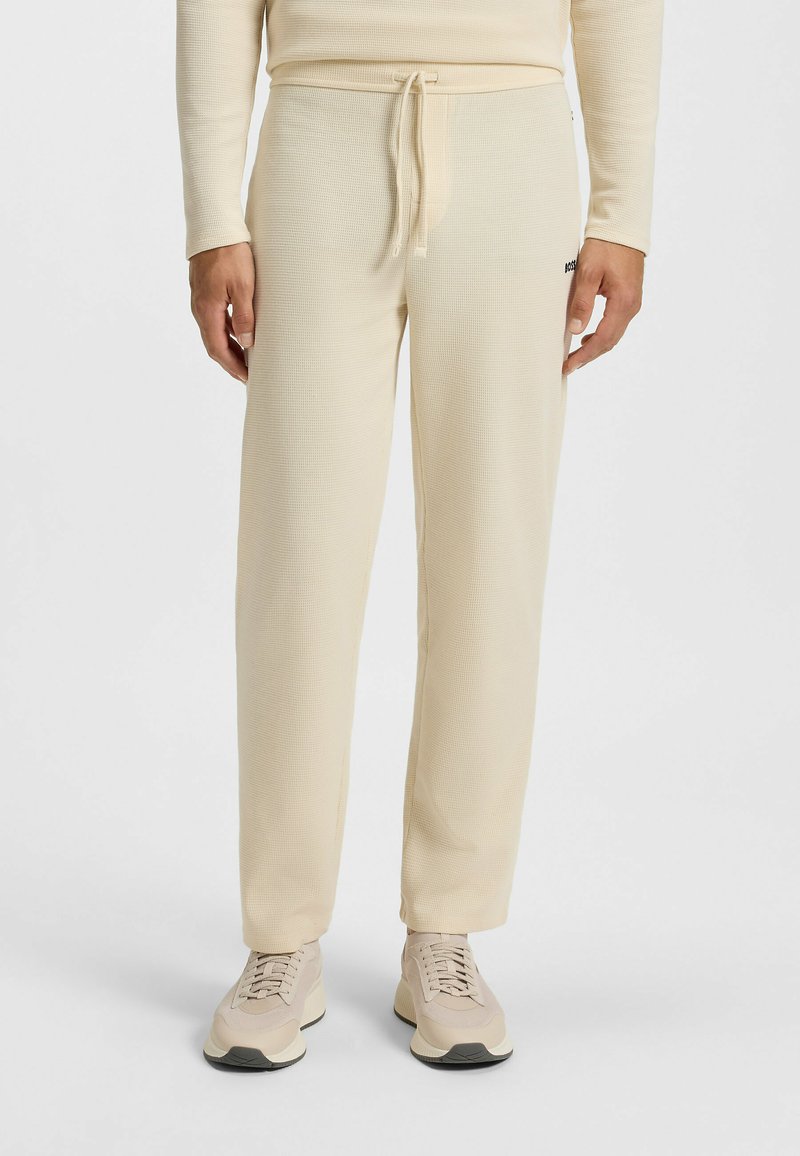 Pantalons texturés en waffle beige avec une taille élastique et un cordon de serrage. Comprend des poches latérales et un petit logo accentué. Portés avec des baskets beiges.