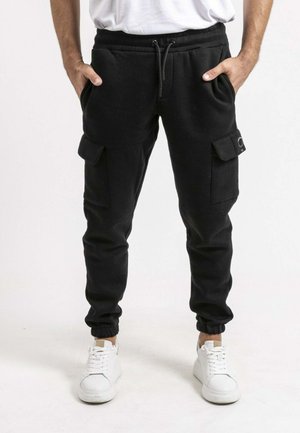 Homme portant un pantalon jogger cargo noir avec poches et taille à cordon, associé à des baskets blanches et une chemise blanche.
