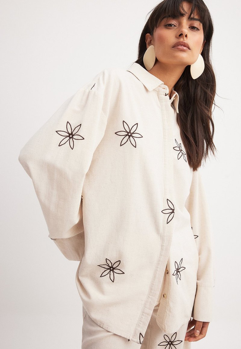 Krémová, dlhá rukávová košeľa vyrobená z textúrovanej látky, s čiernym vyšívaným kvetinovým vzorom na rukávoch a tele, so zapínaním na gombíky.