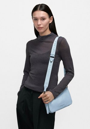 Kobieta o długich ciemnych włosach, ubrana w ciemnoszary top z długim rękawem i czarne spodnie, trzymająca jasnoniebieską torebkę na ramię marki Liebeskind.