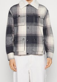 Kraciasta kurtka w kolorach granatowym, szarym i kremowym, z kołnierzem z futerka ekologicznego oraz dwoma przednimi kieszeniami. Wykonana z fakturowanej tkaniny.