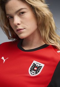 Αθλητική φανέλα σε κόκκινο και μαύρο με κοντά μανίκια, υφασμάτινη με υφή, με λογότυπο της Puma και μια κονκάρδα με θέμα ποδοσφαίρου που ξεχωρίζει.