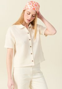 Kortärmad, krämfärgad skjorta med knappar och ett texturerat stickat mönster, med en nedfällbar krage, som kombineras med ljusa byxor.