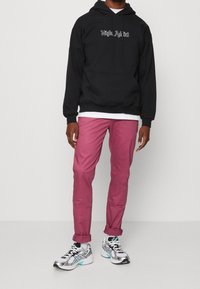 Mężczyzna w czarnej bluzie z kapturem z napisem "Night Addict", białej podkoszulce, różowych spodniaczkach podwiniętych oraz białych srebrnych trampkach, stojący na jasnym tle.