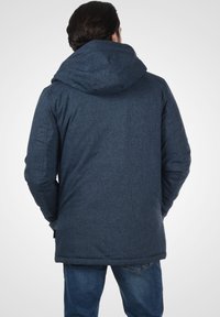 Ciemnoniebieska, textured kurtka z kapturem, o prostym kroju i bocznymi kieszeniami, widziana z tyłu. Casualny design odpowiedni do noszenia na zewnątrz.