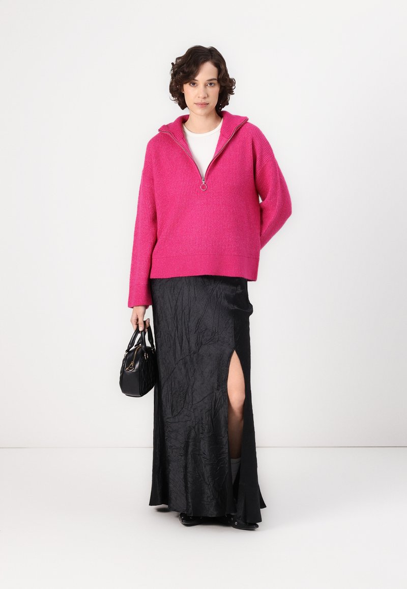 Fuksjowy sweter z dzianiny z zamkiem na szyi, w połączeniu z czarną spódnicą z rozcięciem do uda. Modelka trzyma małą czarną torebkę.