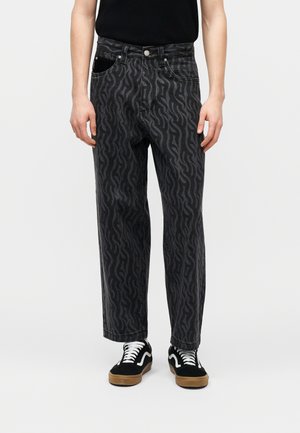 Asmuo dėvi juodus ir tamsiai pilkus banguoto rašto džinsus, juodą marškinėlį ir juodus sportbačius su baltais raišteliais ir želatininėmis padais.