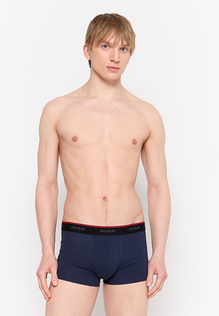 Junger Mann mit blonden Haaren, der dunkelblaue Hugo-Boxershorts trägt und vor einem schlichten weißen Hintergrund steht.