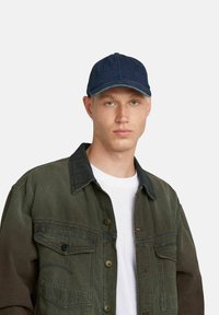Cappello di jeans con corona arrotondata e visiera curvata, di tessuto blu scuro. Modello che indossa una giacca oliva sopra una maglietta bianca.