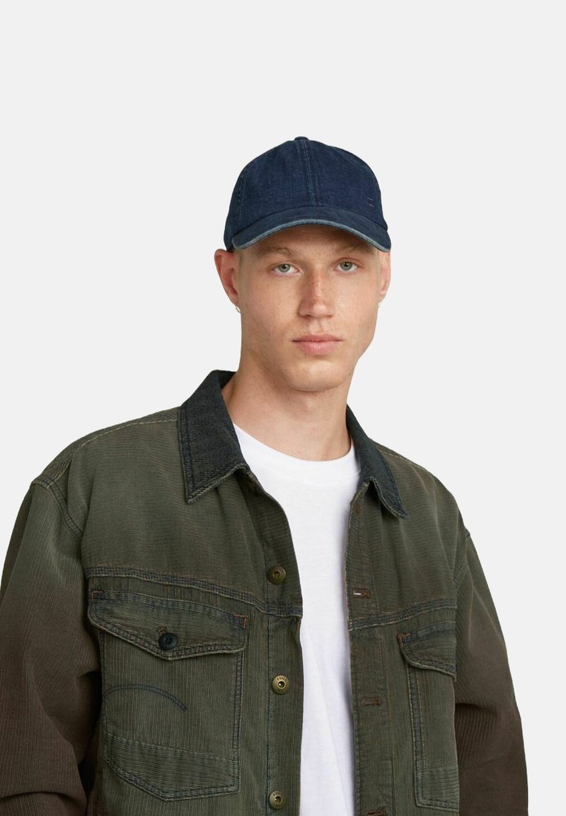 Cappello di jeans con corona arrotondata e visiera curvata, di tessuto blu scuro. Modello che indossa una giacca oliva sopra una maglietta bianca.