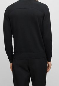 Man wearing a black long-sleeve sweater with ribbed shoulder details and black pants, shown from the back against a gray background.