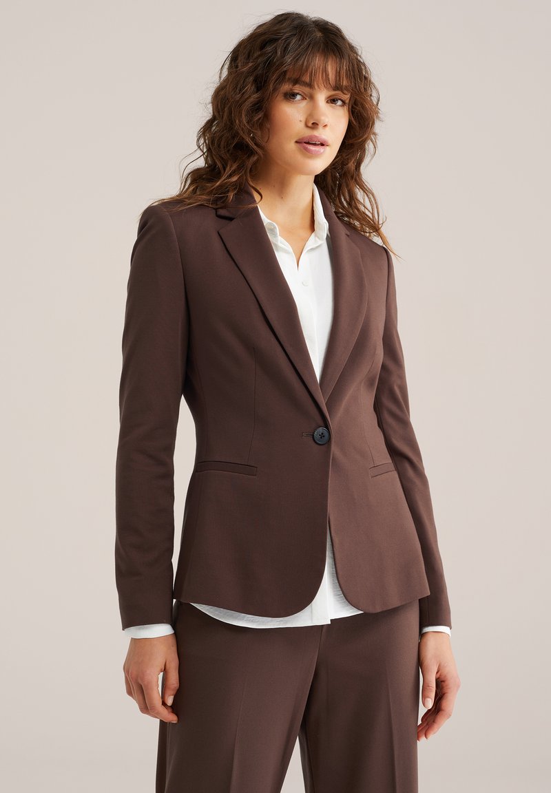 Fiatal nő hullámos barna hajjal, szűk szabású sötétbarna blézert és hozzá illő nadrágot visel, fehér, gombos inget alatt.