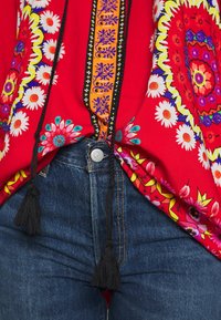 Top floreale rosso con frange nere e motivi vivaci, abbinato a jeans in denim blu con bottone in metallo e cuciture a contrasto.