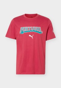 Κόκκινη κοντομάνικη μπλούζα Puma με τη λέξη "PORTUGAL" σε έντονα τιρκουάζ και λευκά γράμματα και το λογότυπο Puma κάτω από αυτά, στο κέντρο μπροστά.