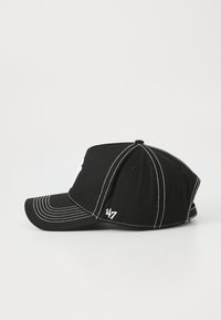Svart baseball-cap med hvite sting og side-logo. Plassert på skakke mot en enkel lys bakgrunn.