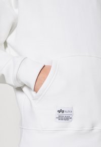 Bela srajca s kapuco in kenguru žepom, opremljena z oznako "Alpha Industries" in mehkim, teksturiranim materialom.