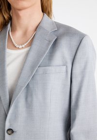 Blazer grigio chiaro con un delicato motivo a quadri, un bottone singolo e una tasca sul petto. Indossato sopra una maglietta a righe e accessoriato con una collana di perle.