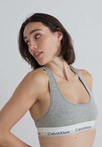 Γκρι αθλητικό σουτιέν με στρογγυλή λαιμόκοψη και σχέδιο racerback. Διαθέτει λευκή ζώνη με το λογότυπο "Calvin Klein". Μαλακή υφή υφάσματος.