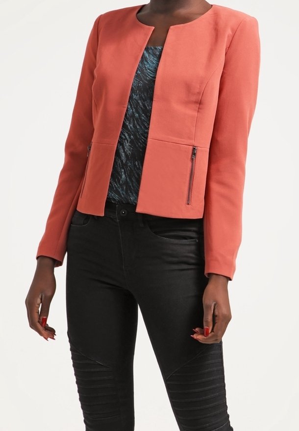 Blazer corail avec un col arrondi, une coupe structurée et des poches zippées, associé à un haut bleu à motifs et un pantalon noir ajusté.