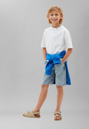 Bambino con capelli biondi che indossa una t-shirt bianca Calvin Klein, felpa blu legata in vita, pantaloncini di jeans e sandali beige, sorridente.