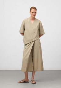 Completo in cotone leggero di colore beige, composto da una maglia avvolgente con maniche corte e pantaloni a gamba larga. Sandali piatti completano il look.