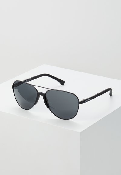 Ochelari de soare aviator negri cu lentile gri, cadru metalic subțire și brațe din plastic texturat negru. Afișați pe o suprafață albă.