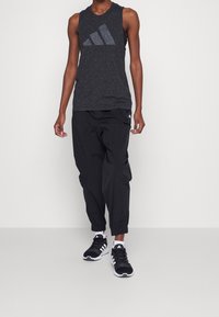 Canottiera atletica nera senza maniche con texture maculata e logo grigio, abbinata a pantaloni neri larghi e scarpe da ginnastica nere.