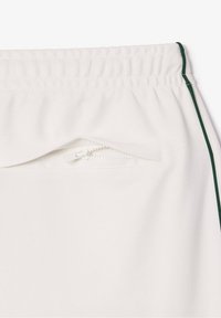 Pantalon de sport blanc avec une surface texturée, doté d'une poche zippée et d'accents verts sur les côtés. Taille élastique pour un ajustement sécurisé.