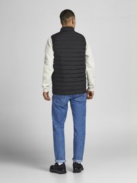 Zwarte puffer vest met verticale stikpatronen, gedragen over een crèmekleurig sweatshirt, gecombineerd met lichtblauwe opgerolde jeans en zwarte sneakers.