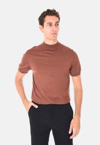 Uomo che indossa una camicia marrone a maniche corte e pantaloni neri, in piedi con una mano in tasca, mentre guarda di lato su uno sfondo neutro.