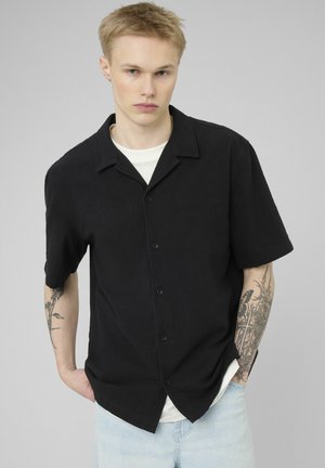 Jeune homme aux cheveux blonds courts portant une chemise noire à manches courtes boutonnée sur un t-shirt blanc, avec des tatouages visibles sur les deux avant-bras.